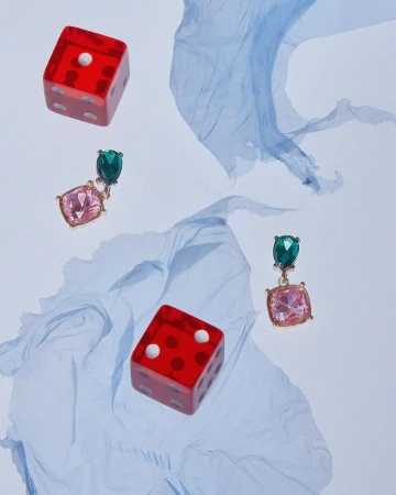 Studio shot featuring red dice, gemstone earrings with green and pink stones, and light blue crumpled fabric on a white backdrop.