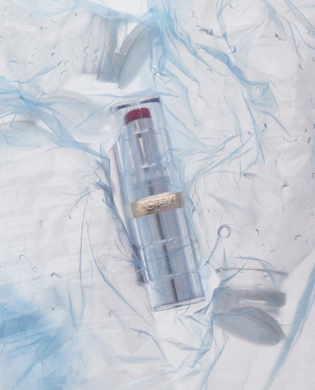 Red lipstick tube by L'Oreal in a translucent container displayed on light blue crinkled plastic sheet with subtle water droplets.