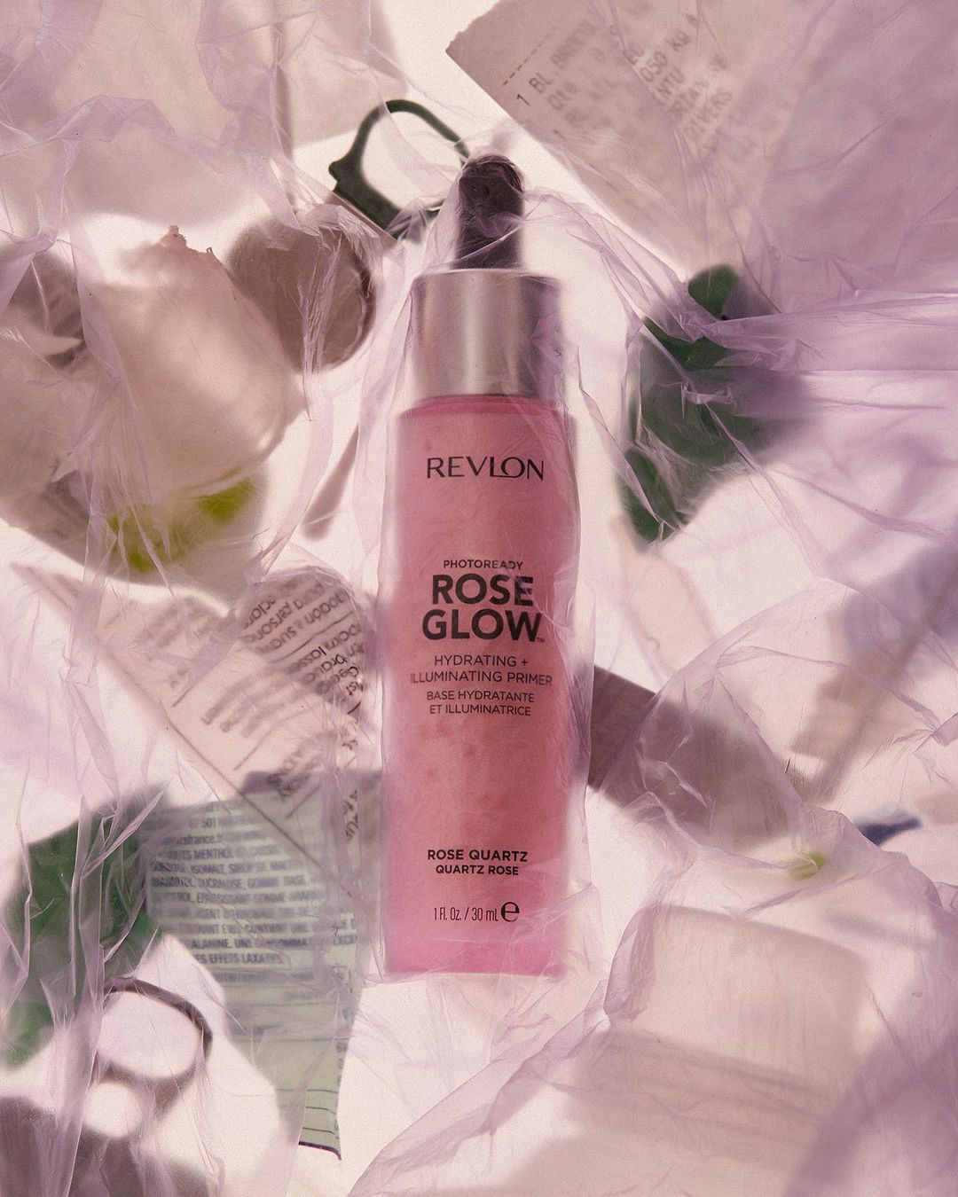 Revlon Rose Glow primer bottle is displayed against a background of crumpled receipts and packaging in a softly lit, artistic still life.