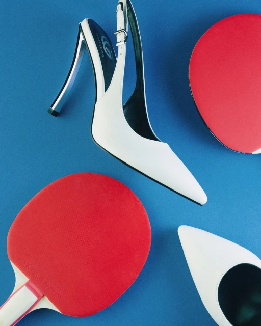 Elevated view of a white slingback heel positioned between two red ping pong paddles on a vibrant blue background.