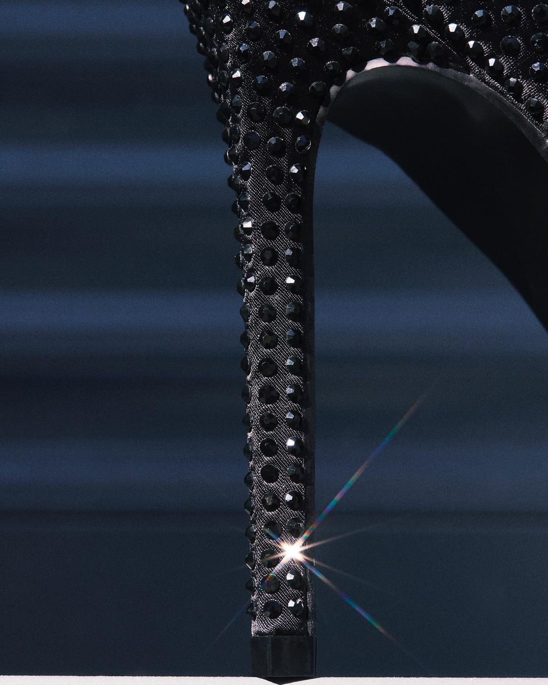 Close-up of black stiletto high heel with rhinestones. Focus on the heel and platform against blurred dark blue background.