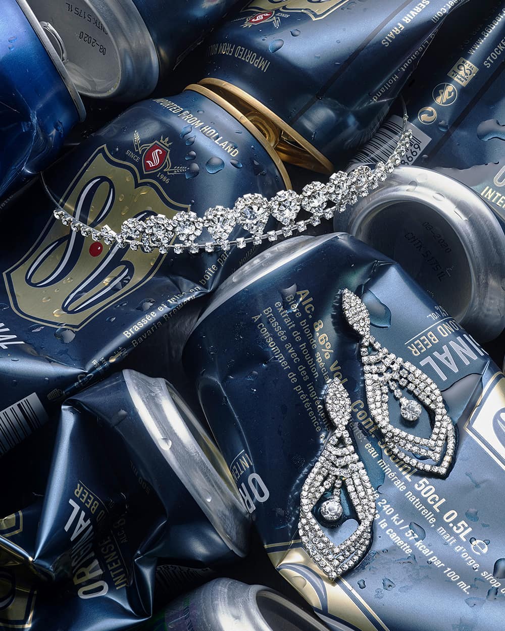 Sparkling earrings and tiara rest atop crushed blue beer cans. Elegant jewelry contrasts with the casual beverage containers.