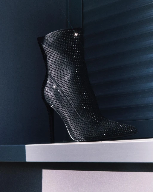 Black high-heeled ankle boot, covered in rhinestones, positioned on a light grey surface against a dark blue paneled background.