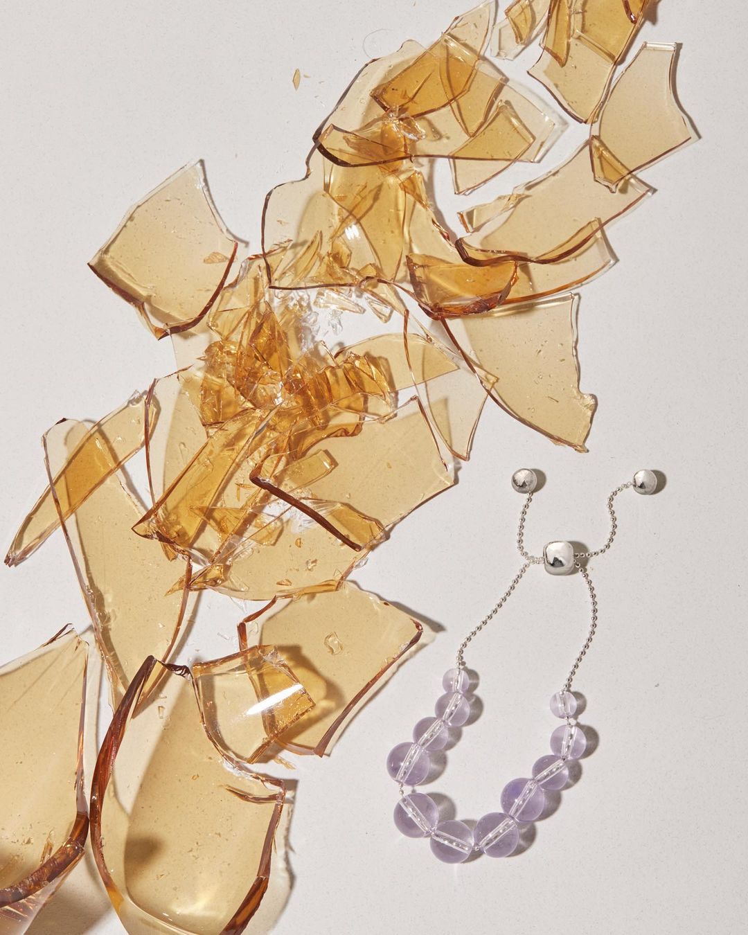 Still life shot featuring a bracelet with lavender beads lying near shattered amber-colored glass fragments on a white background.