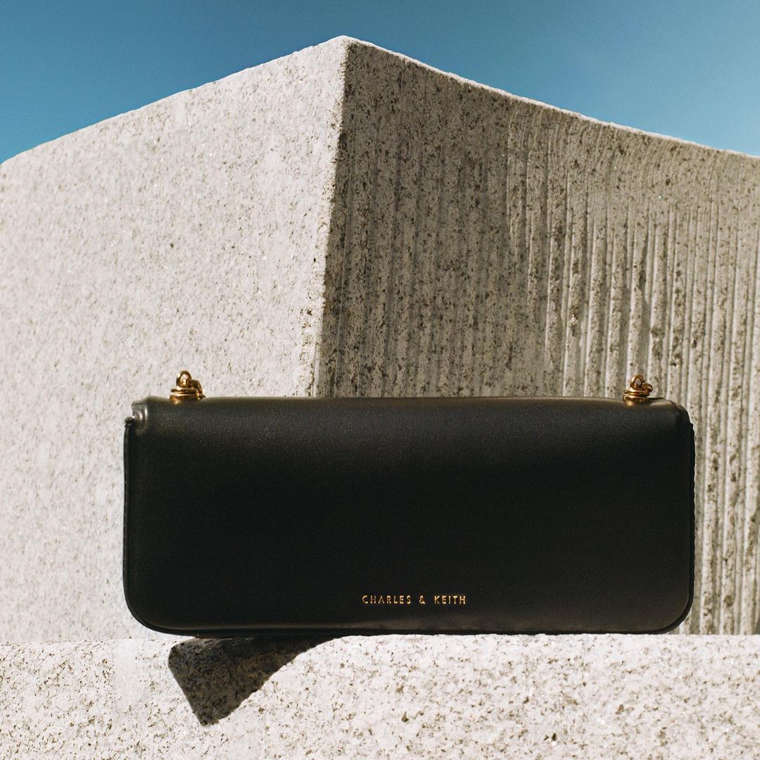 Black leather shoulder bag with gold hardware resting on textured grey concrete blocks against a clear blue sky.