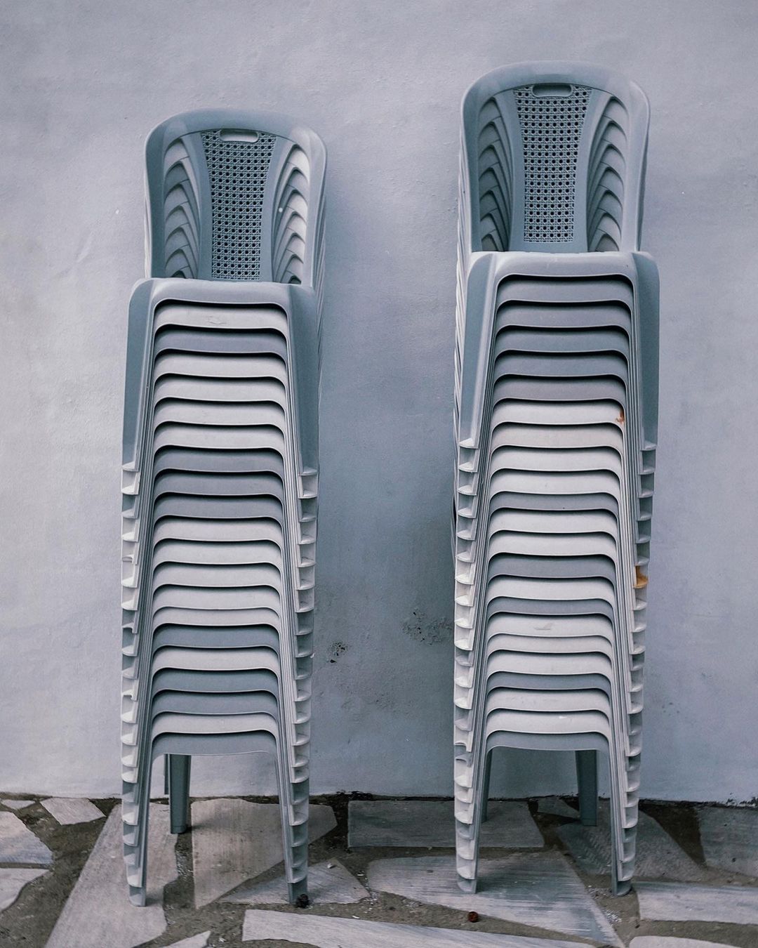 Stacked gray plastic chairs with perforated backs against a weathered gray wall and tiled floor in an indoor or outdoor setting.