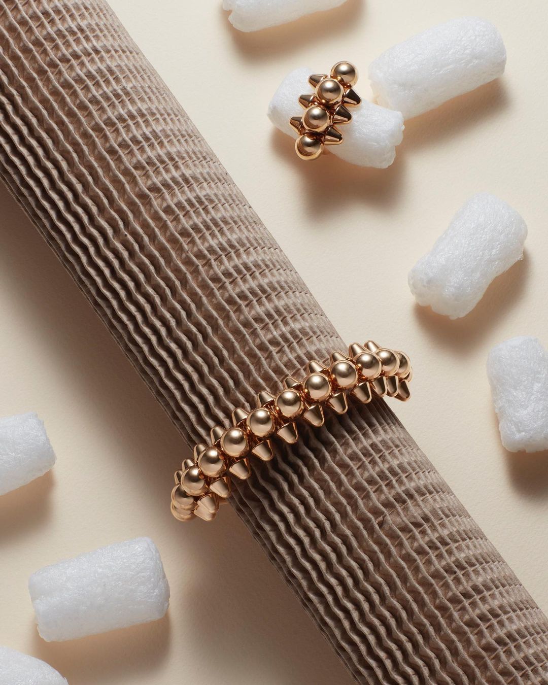 Elevated product shot of a gold-toned beaded bracelet and ring on a beige corrugated tube, surrounded by white packing peanuts.