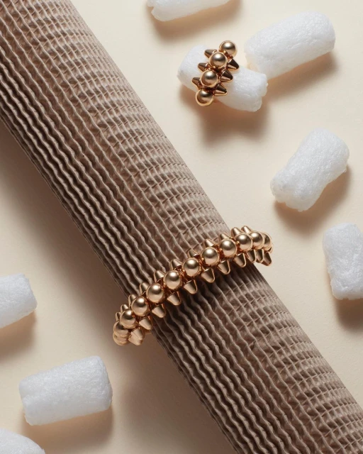 Elevated product shot of a gold-toned beaded bracelet and ring on a beige corrugated tube, surrounded by white packing peanuts.