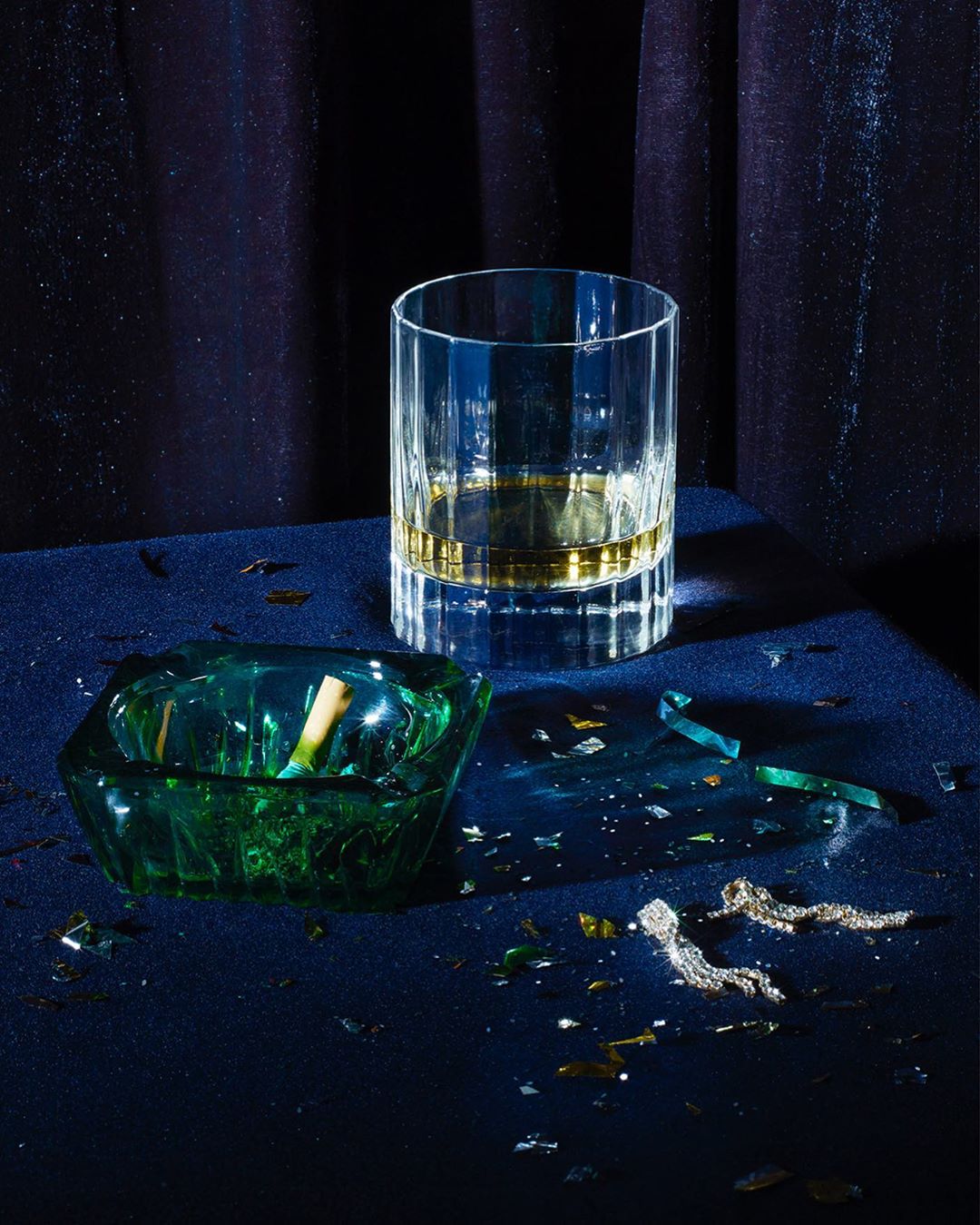 Studio shot of a glass of whiskey and a green ashtray with a cigarette butt, against a blue velvet curtain, and broken glass.