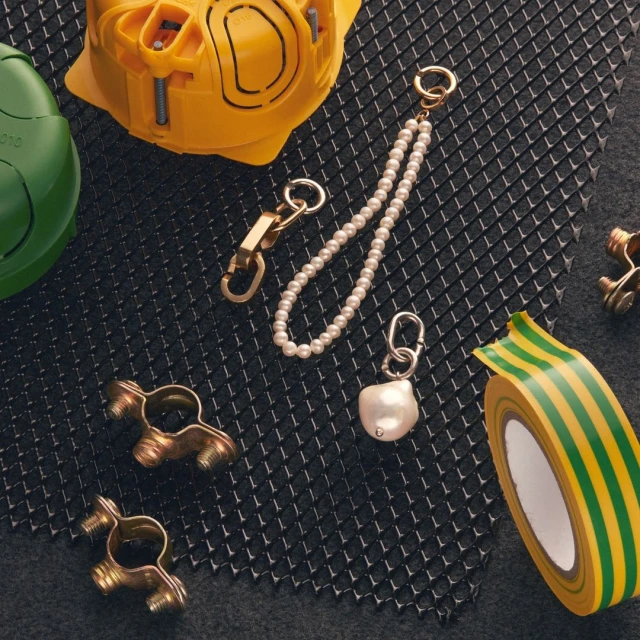 Still life of hardware and jewelry arranged on a black grid surface, featuring pearls, metal fasteners, and yellow tape.