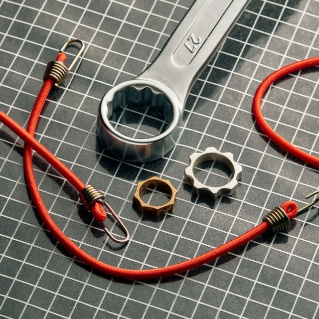 Close-up still life of a silver wrench, two metal rings, and a red bungee cord on a dark gray surface with a grid pattern.