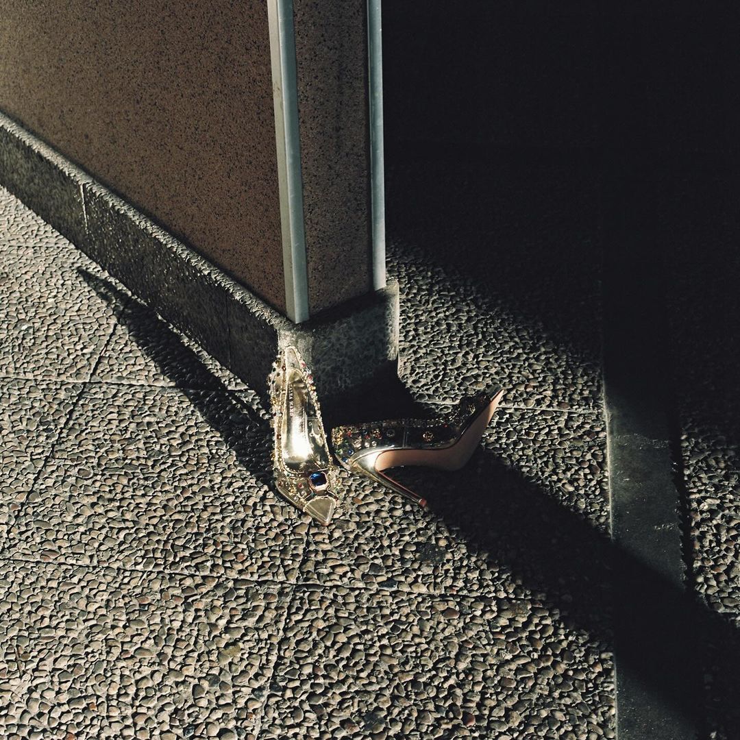 High-heeled shoes rest on a stone-tiled surface, illuminated by a combination of natural light and dark shadows near a beige wall.