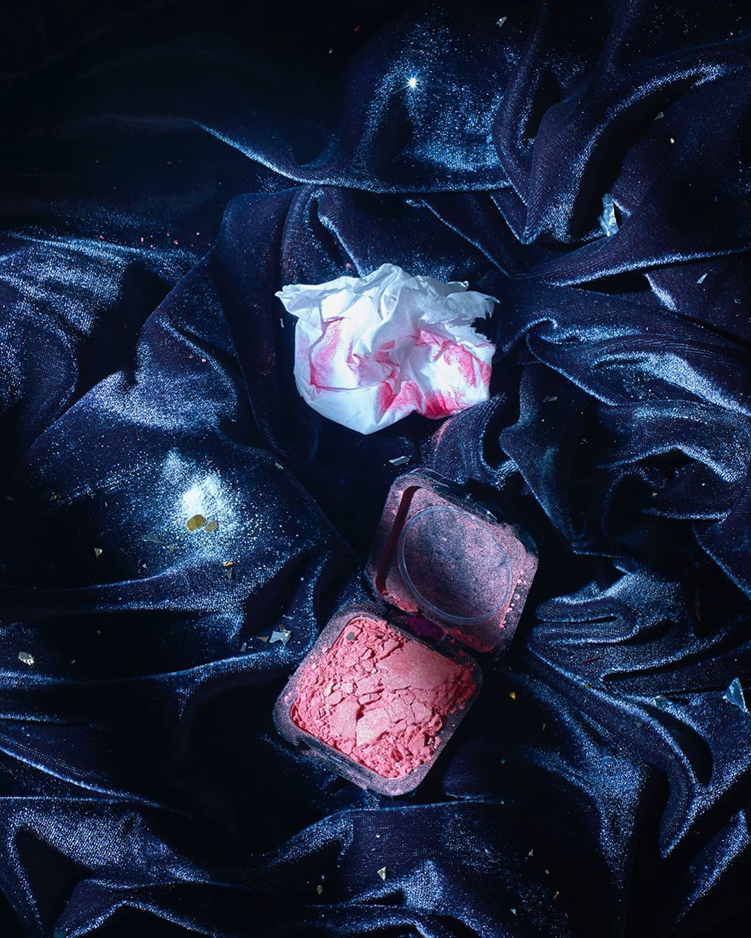 Close-up studio shot of dark blue velvet fabric with an open pink blush compact and a crumpled tissue with red lipstick marks.
