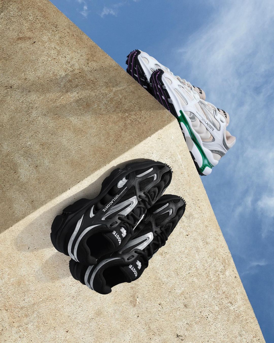 Two pairs of Lacoste sneakers, one black and one white, are displayed against a concrete ledge with a background view of the sky.