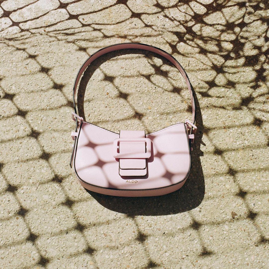 A light pink shoulder bag with a prominent buckle rests on a textured stone ground under a patterned shadow.