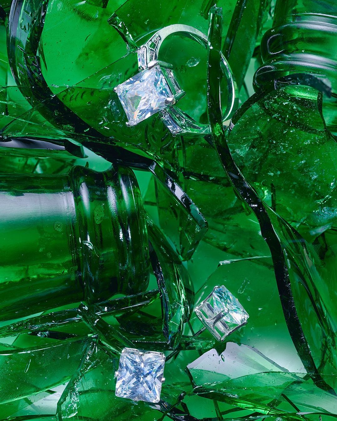 A close-up of a ring and earring set featuring princess-cut gemstones surrounded by green broken glass, shot with studio lighting.