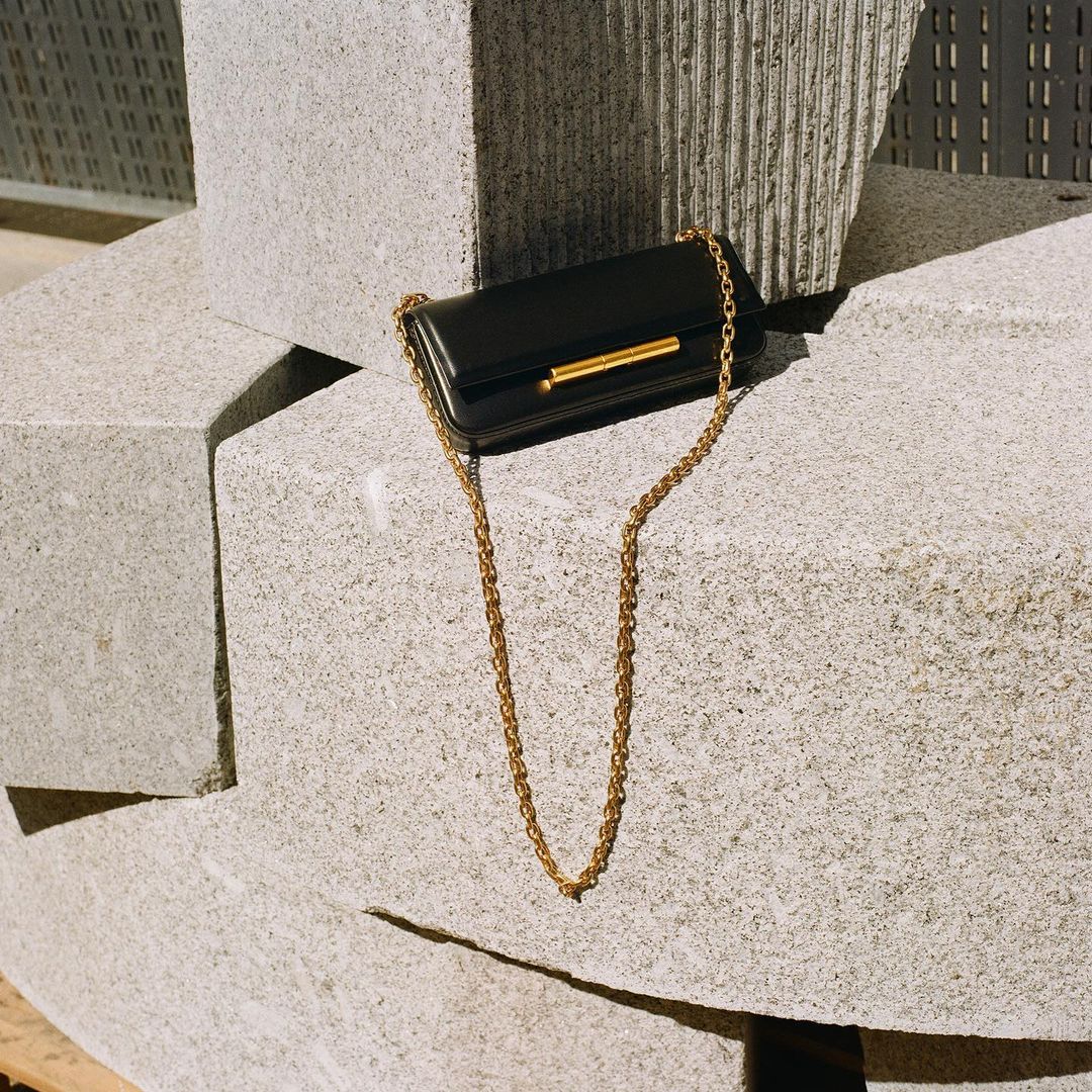 Elegant black purse with a gold chain strap, displayed against a backdrop of gray concrete blocks on a sunny day.
