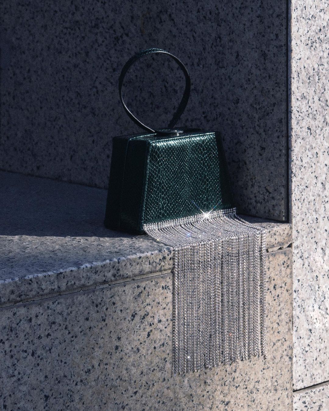 Emerald green structured handbag with a circular handle and a long, sparkling silver fringe, showcased against a granite stone backdrop.