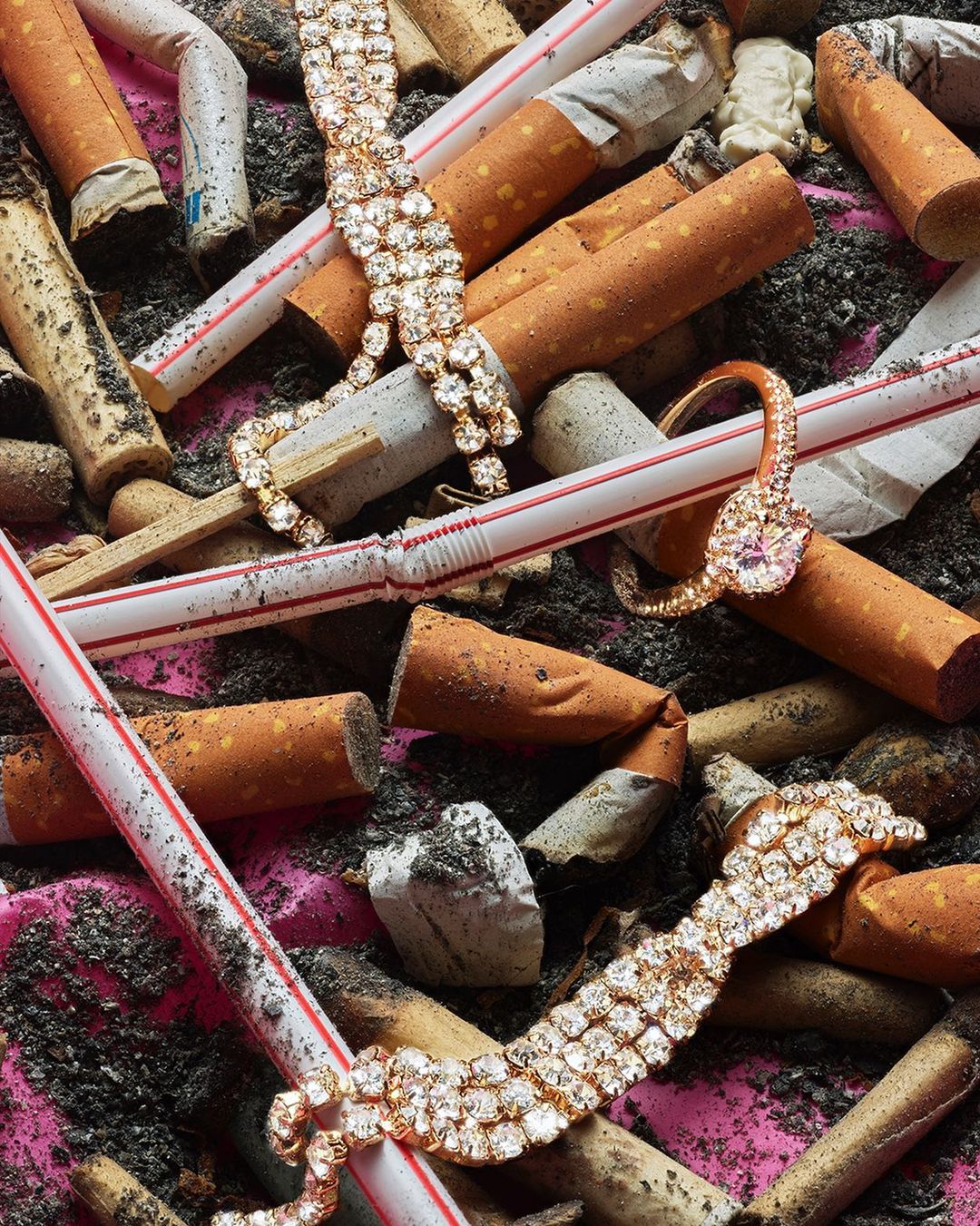 Scattered diamond bracelets and a ring amidst discarded cigarette butts and drinking straws on a wrinkled pink surface.
