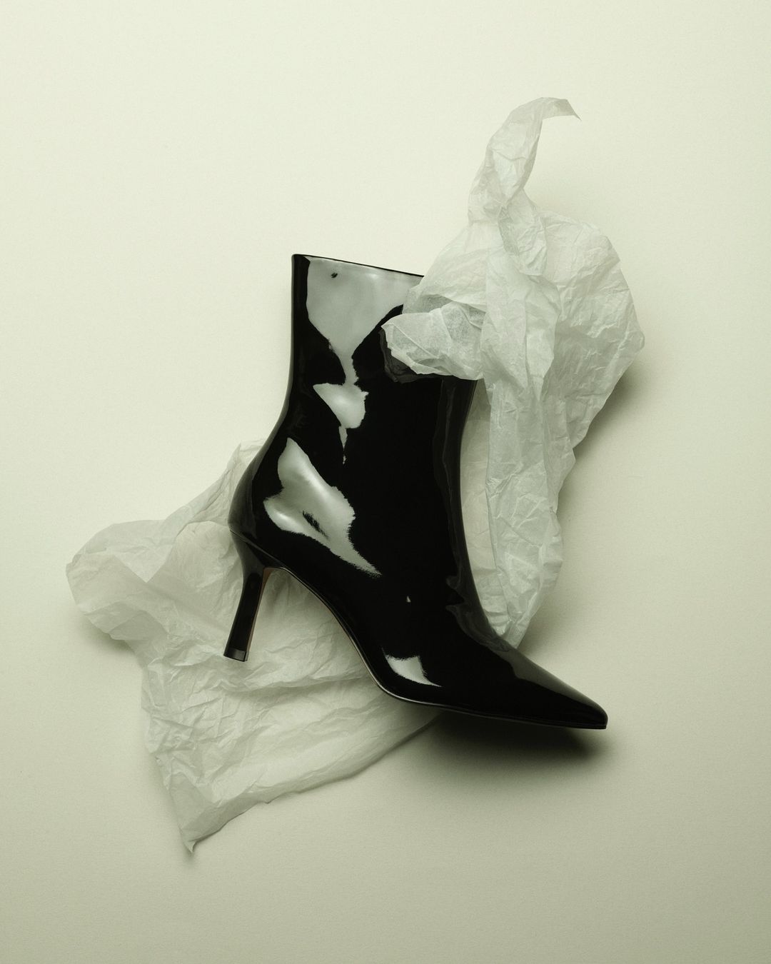 High-gloss black patent leather ankle boot with a pointed toe, displayed with crumpled white tissue paper on an off-white surface.