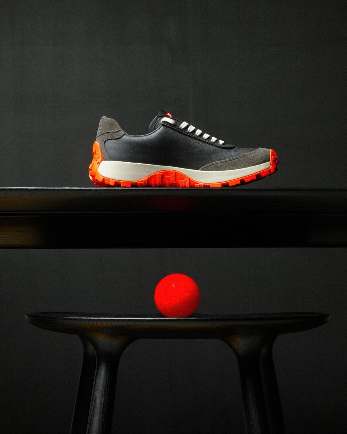 Studio shot of a black leather and gray suede sneaker with orange accents, placed on a black table with a red ball underneath.