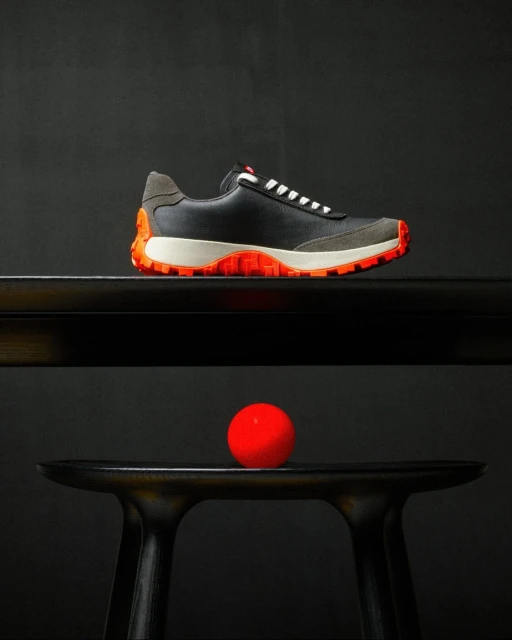 Studio shot of a black leather and gray suede sneaker with orange accents, placed on a black table with a red ball underneath.