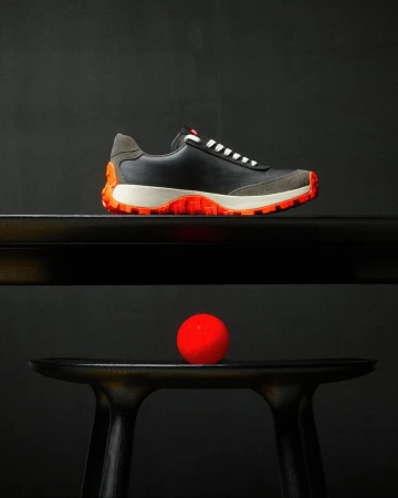 Studio shot of a black leather and gray suede sneaker with orange accents, placed on a black table with a red ball underneath.