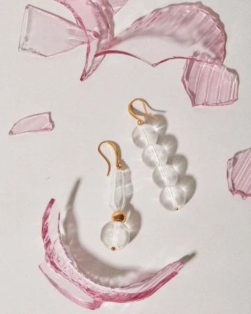 Clear glass bead earrings with gold hooks displayed on a light surface surrounded by broken translucent pink glass shards.