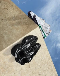 Two pairs of sneakers resting on an angled concrete wall against a bright blue sky with wispy white clouds.