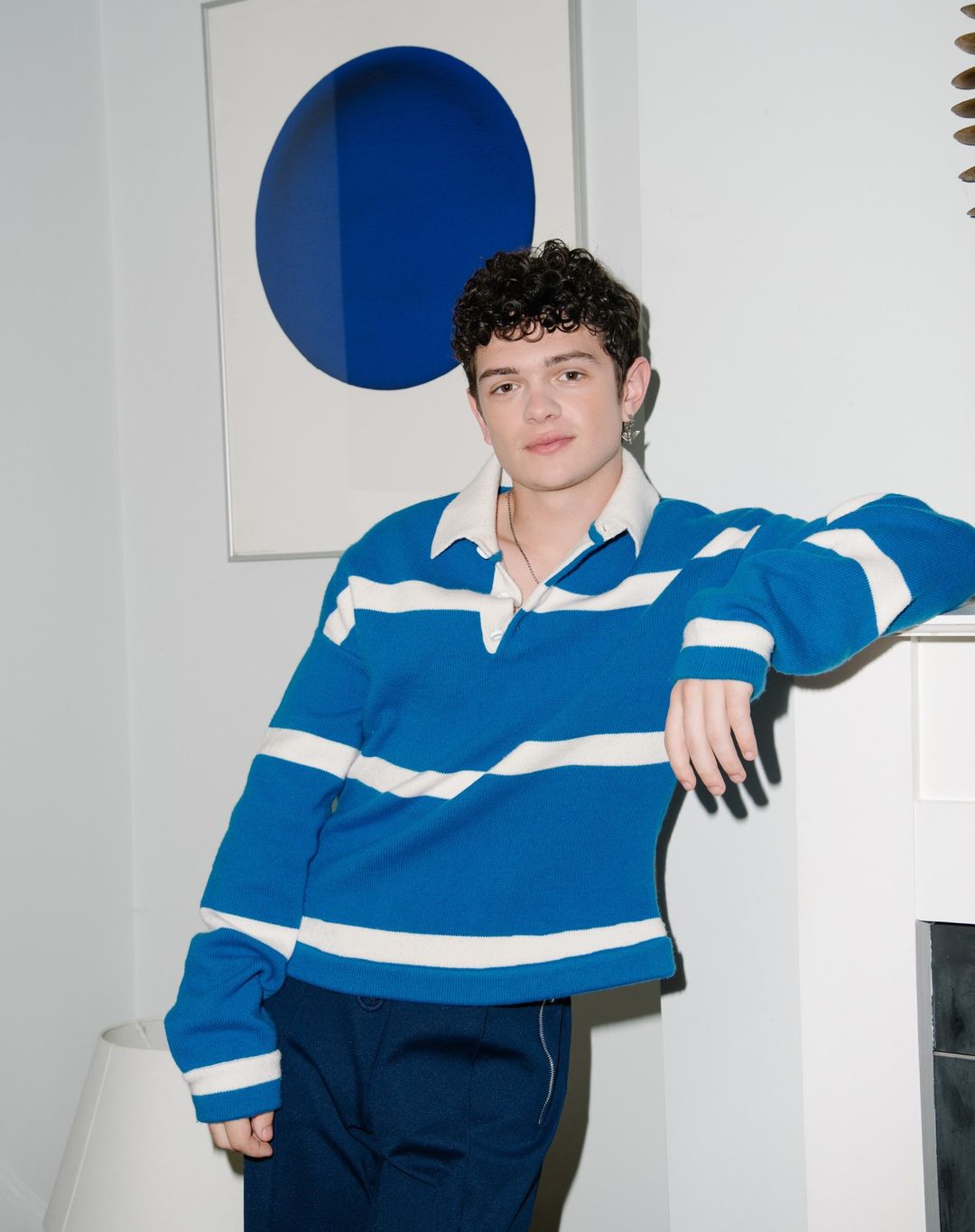 Portrait of a young adult male in a blue and white rugby shirt, posing indoors with blue circle art and fireplace backdrop.