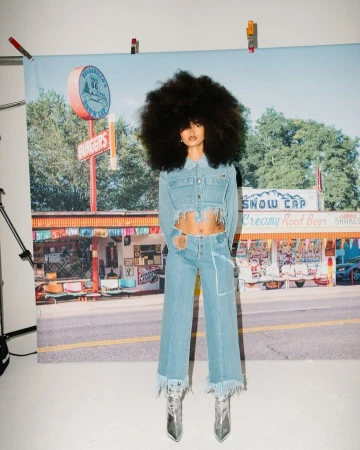 Model with large afro hairstyle wearing a cropped denim jacket and matching jeans in front of a diner backdrop.