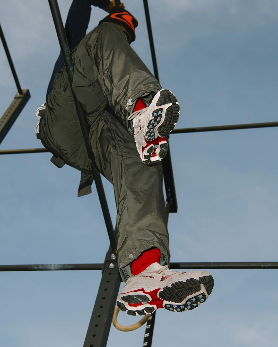 Person climbing monkey bars wearing athletic shoes. A low-angle perspective captures the gray metal bars against a bright blue sky backdrop.