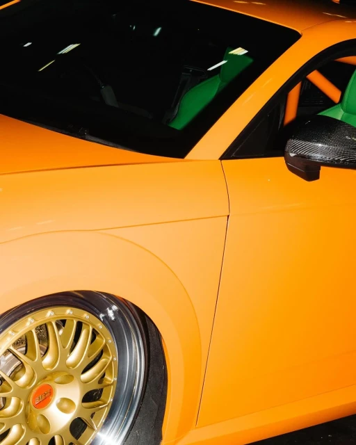 Close-up of a bright orange Audi TT featuring gold rims and carbon fiber mirror. Car enthusiasts will appreciate this custom vehicle.