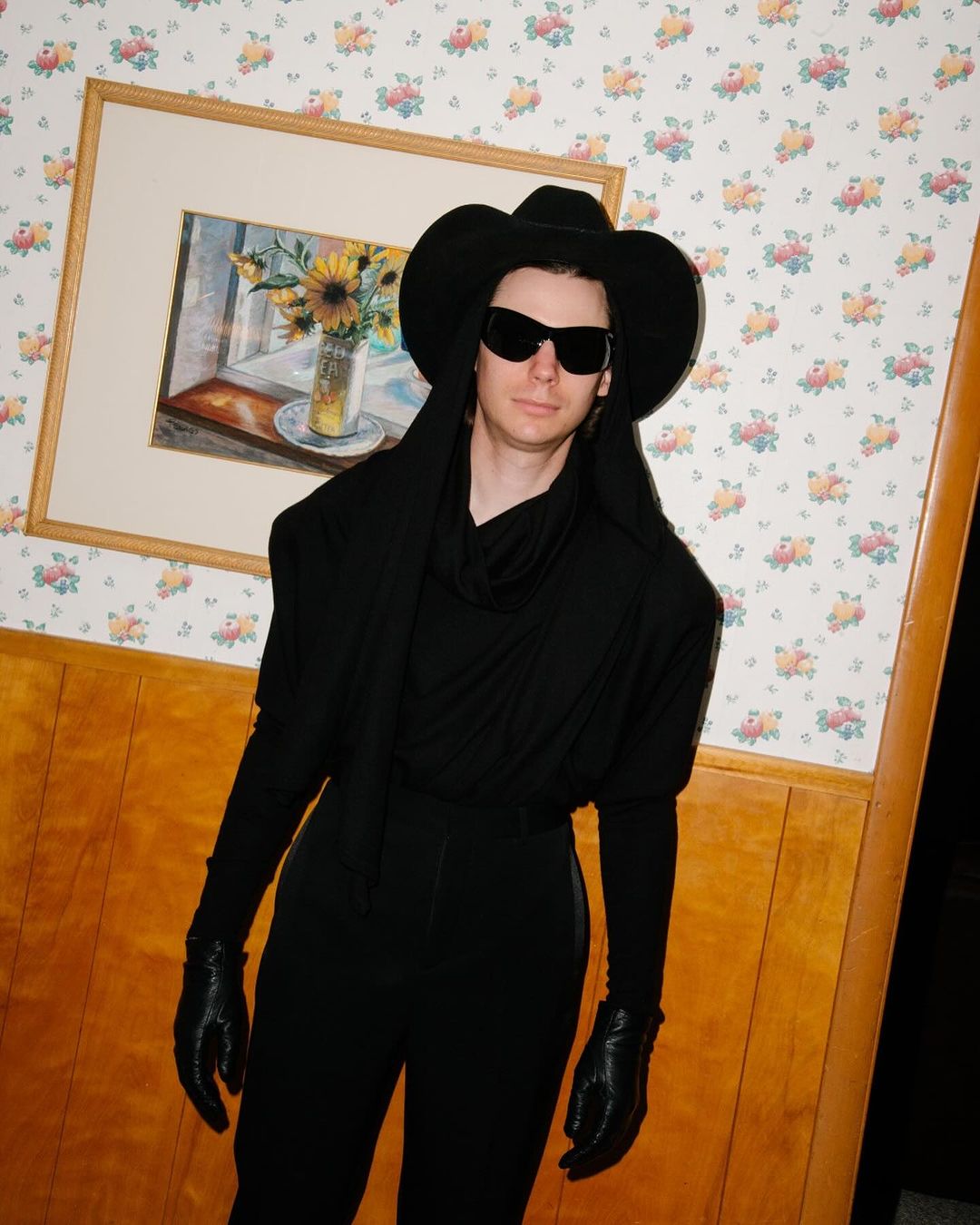 Person posing in all-black outfit with cowboy hat and sunglasses, standing indoors against floral wallpaper and framed art.