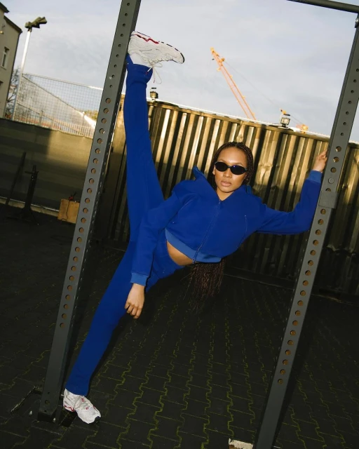 An adult female does a split pose on a metal structure outdoors, wearing a blue tracksuit and sunglasses against an urban backdrop.