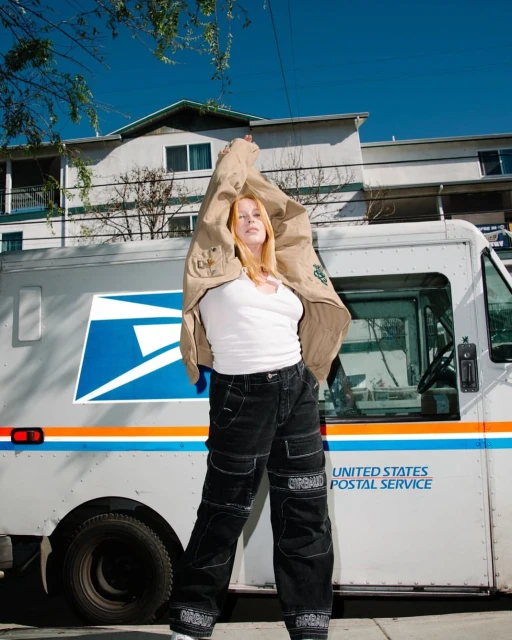 Adult female model wearing dark pants with a white top standing in front of a white United States Postal Service delivery truck.