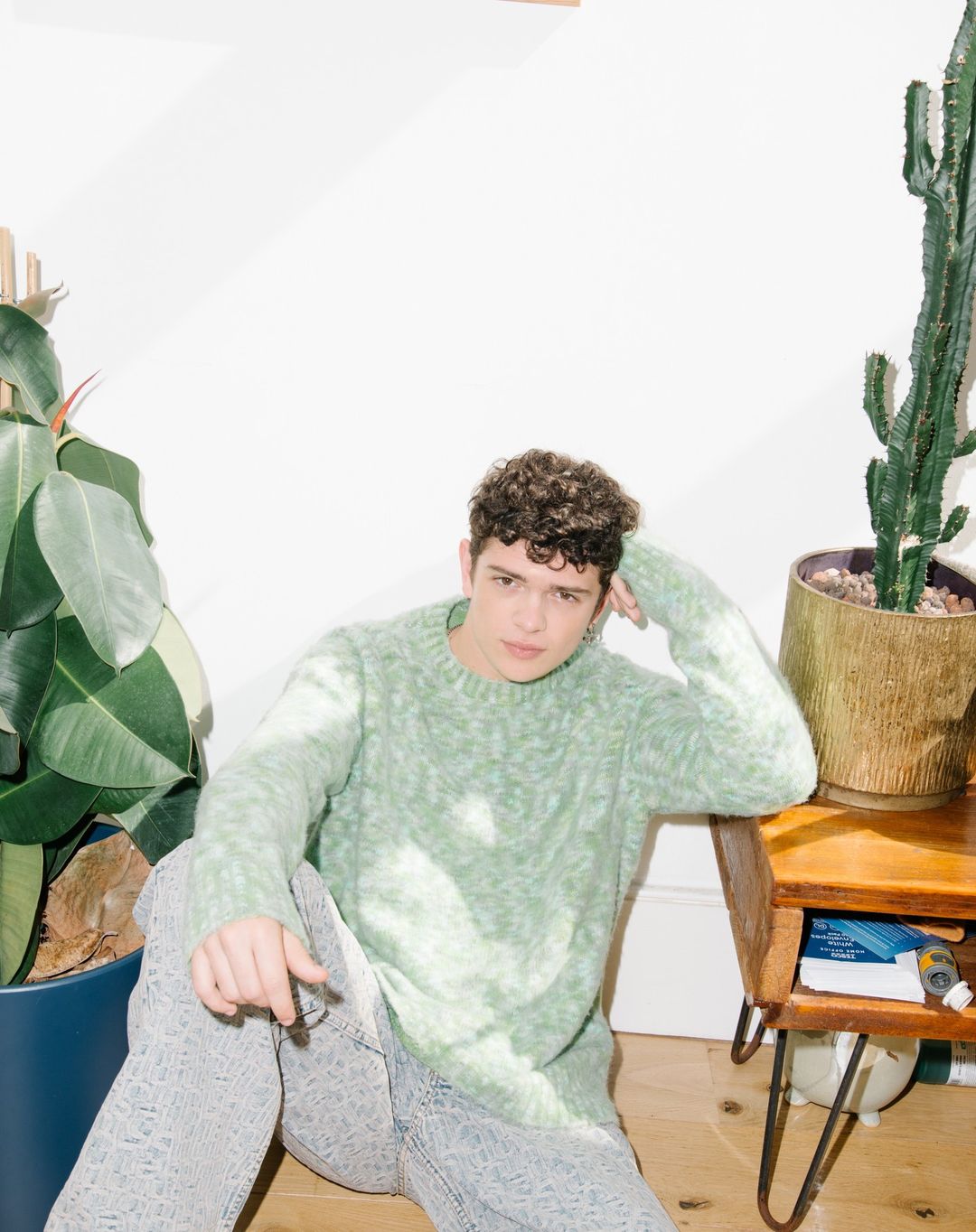 Young adult man in green sweater and patterned jeans sitting indoors next to potted plants, styled with casual and candid pose.