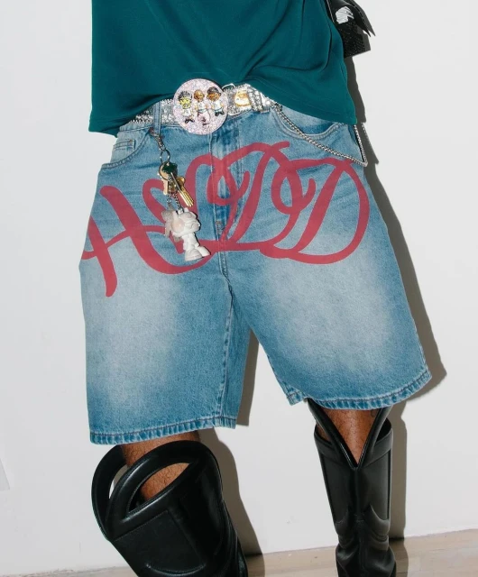 Cropped studio shot of a person wearing denim shorts with red graffiti print, accessorized with a belt, keys, and black boots.