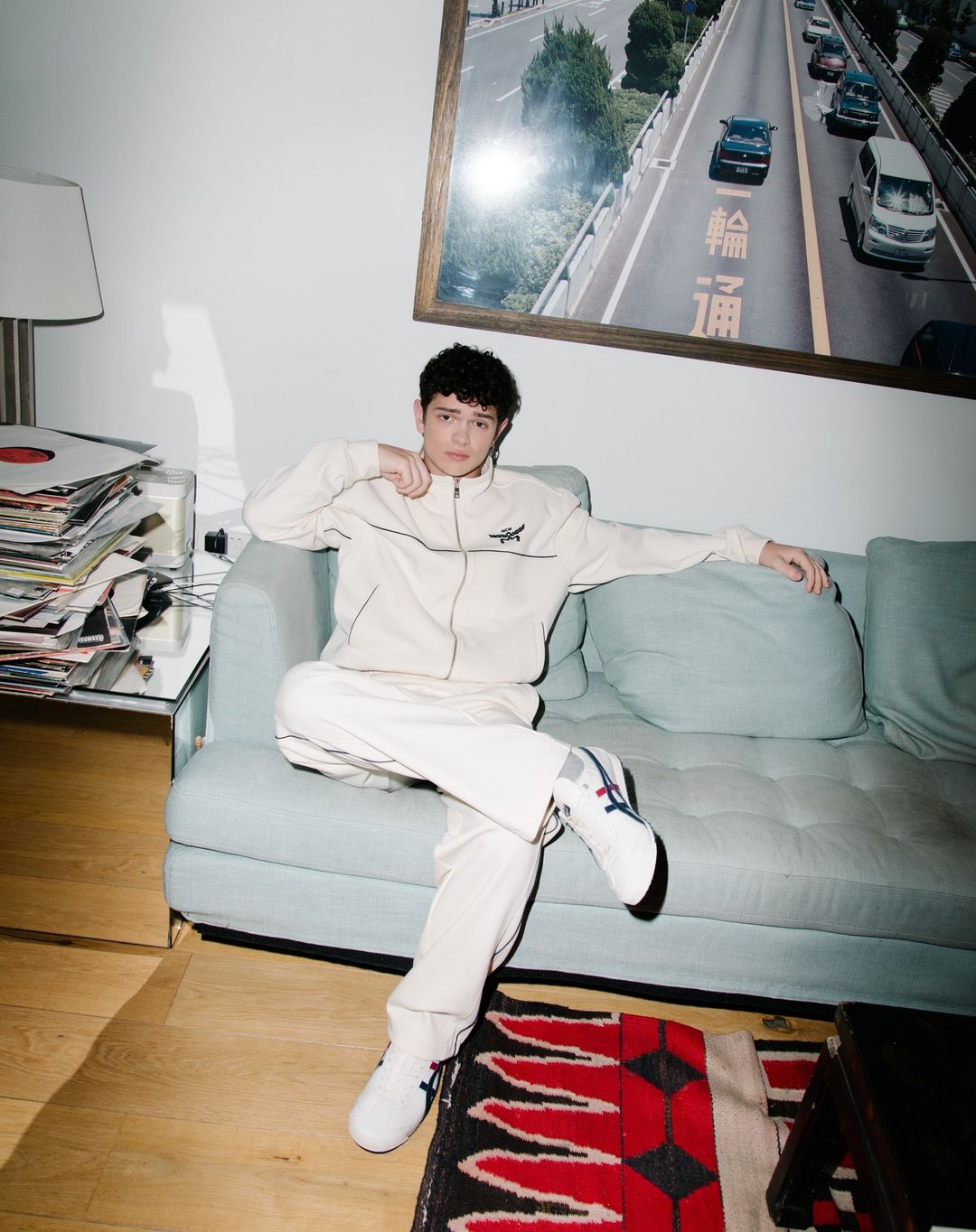 Young adult poses in a beige tracksuit on a blue sofa in a brightly lit interior, featuring records, rug, and a road photo.