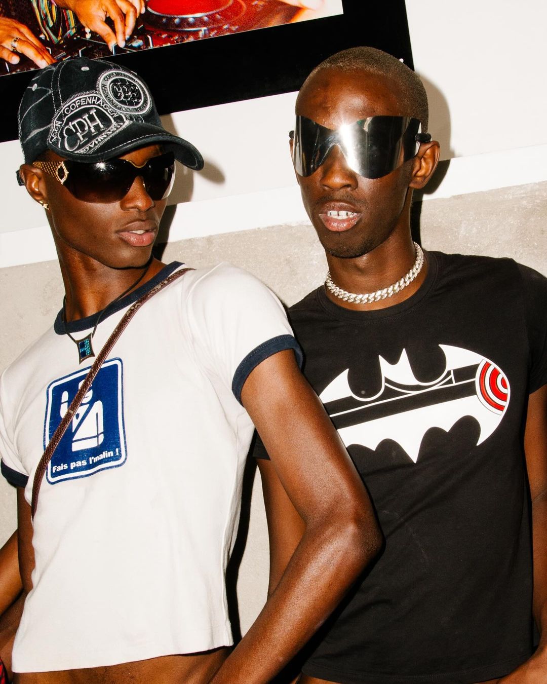 Two adult male fashion models with dark skin, wearing sunglasses and t-shirts, pose indoors in front of a white wall with artwork.