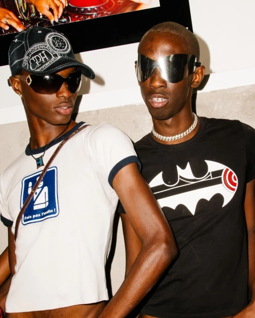 Two adult male fashion models with dark skin, wearing sunglasses and t-shirts, pose indoors in front of a white wall with artwork.