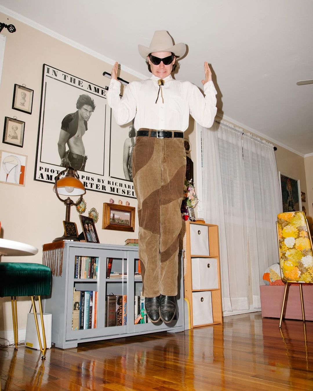 An adult wearing a cowboy hat and brown pants floats in the air inside a room, which has a grey bookshelf and yellow patterned chair.