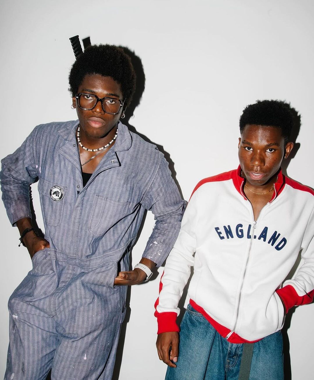 Two stylish young men pose indoors against a white wall. One wears blue coveralls and glasses; the other, a white jacket and jeans.
