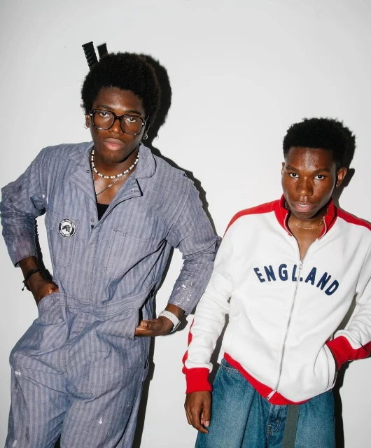 Two stylish young men pose indoors against a white wall. One wears blue coveralls and glasses; the other, a white jacket and jeans.