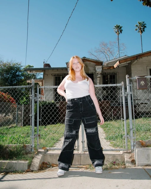 Full-length portrait of a red-haired woman in baggy black pants and a white tank top in front of a chainlink fence and a home.