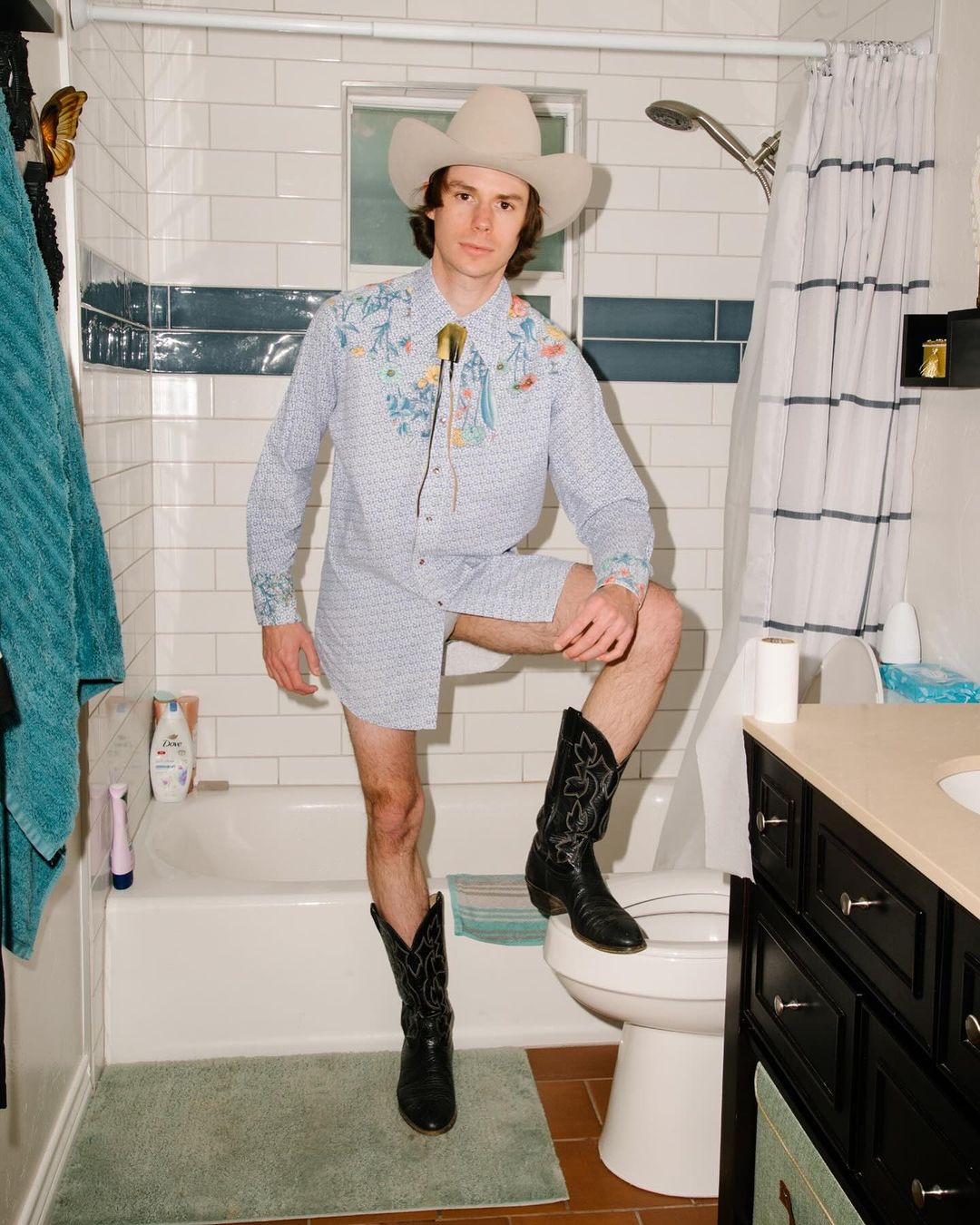 Man poses in bathroom wearing cowboy hat, shirt, shorts, and boots while sitting on toilet with one leg on bathtub edge.