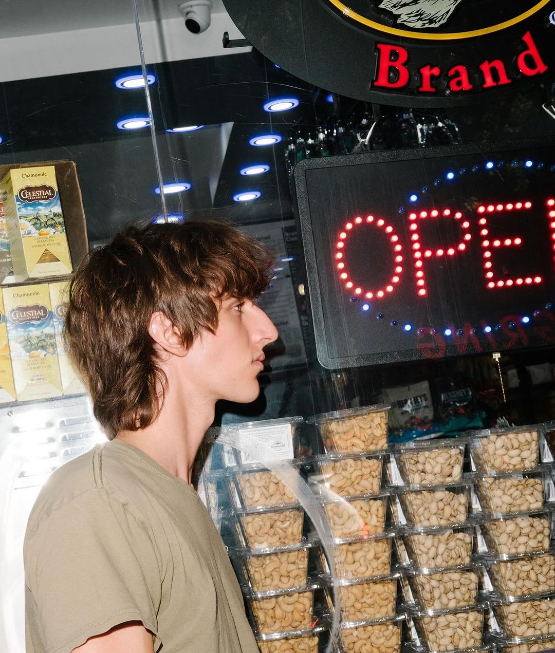 Profile view of a young adult in a green shirt, standing near shelves stocked with pistachios and in front of an illuminated 'OPEN' sign.