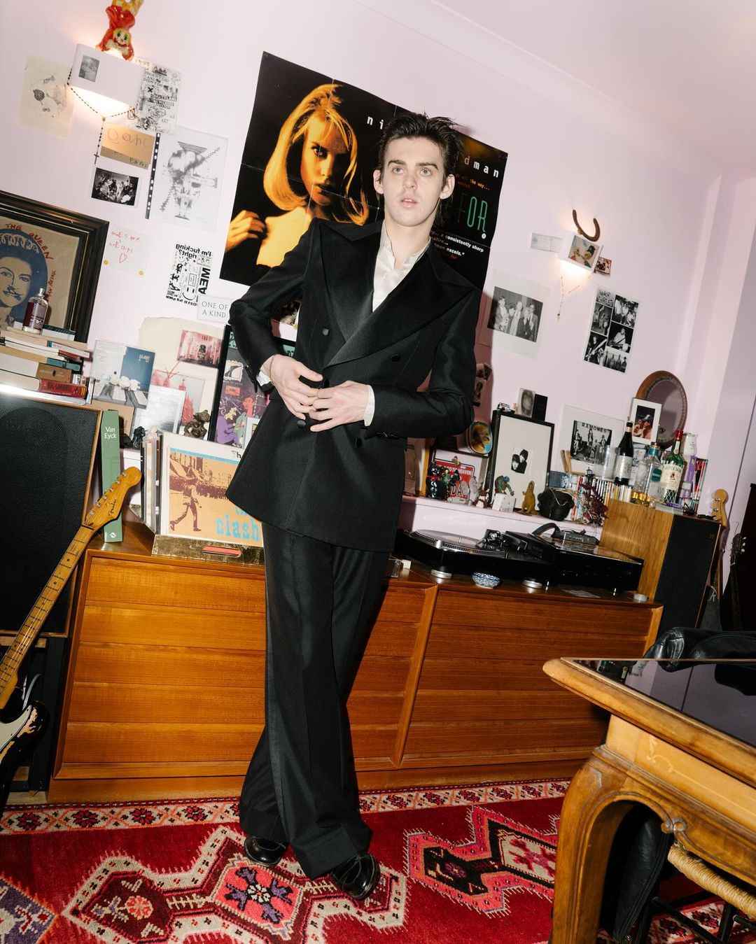 Stylish young man in a black suit poses in a room with posters, records, and a guitar, captured in a full shot with indoor lighting.