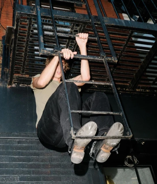 A young adult climbs a metal fire escape ladder outdoors, wearing jeans and boots in an urban setting, captured from a low angle.