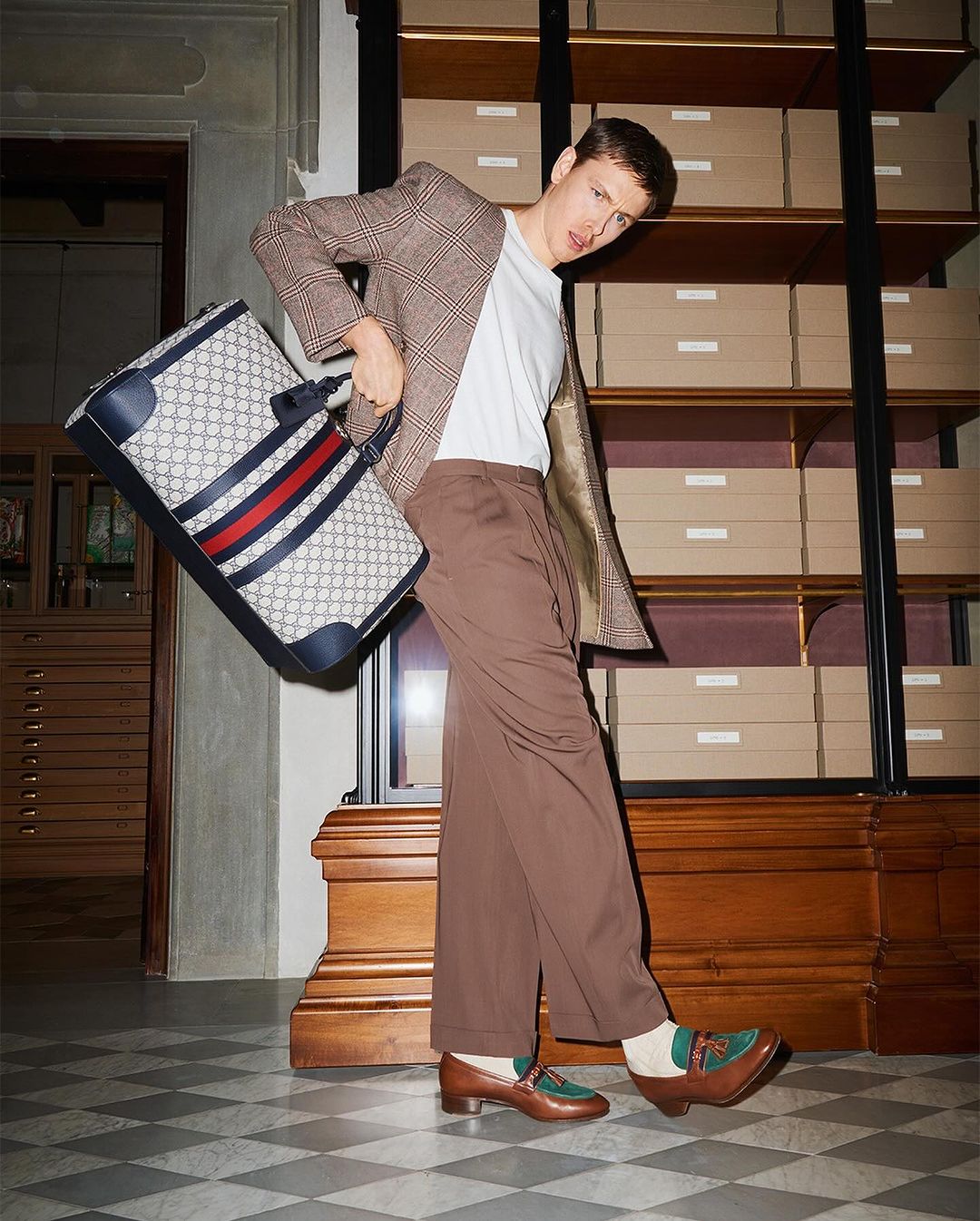 Man in retro fashion holds Gucci travel bag in vintage interior with shelves. Brown plaid jacket, trousers, and loafers.